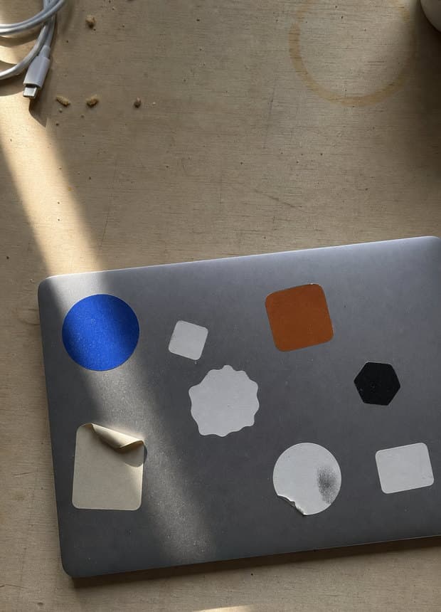 A closed laptop on a wooden desk, its lid covered in plain unlabelled stickers, one peeling at the corner.