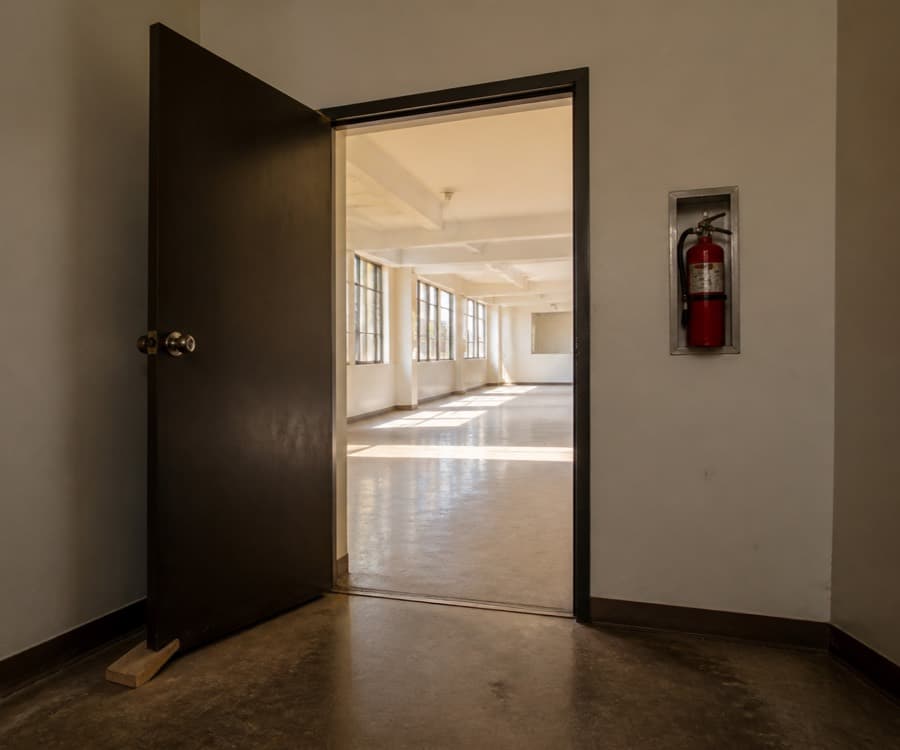 A heavy door propped open with a wooden wedge onto a bright empty room with windows down one side and clear sightlines across the floor.