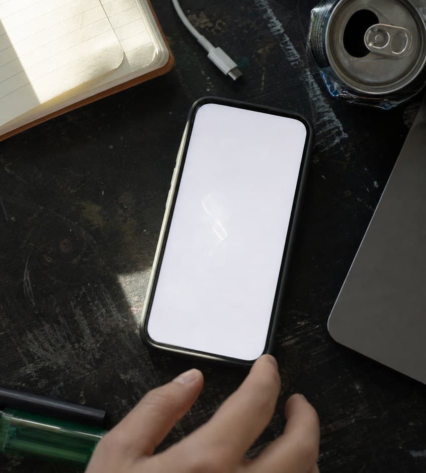 A phone face up on a scratched desk beside a notebook and a laptop, a hand reaching in to pick it up.