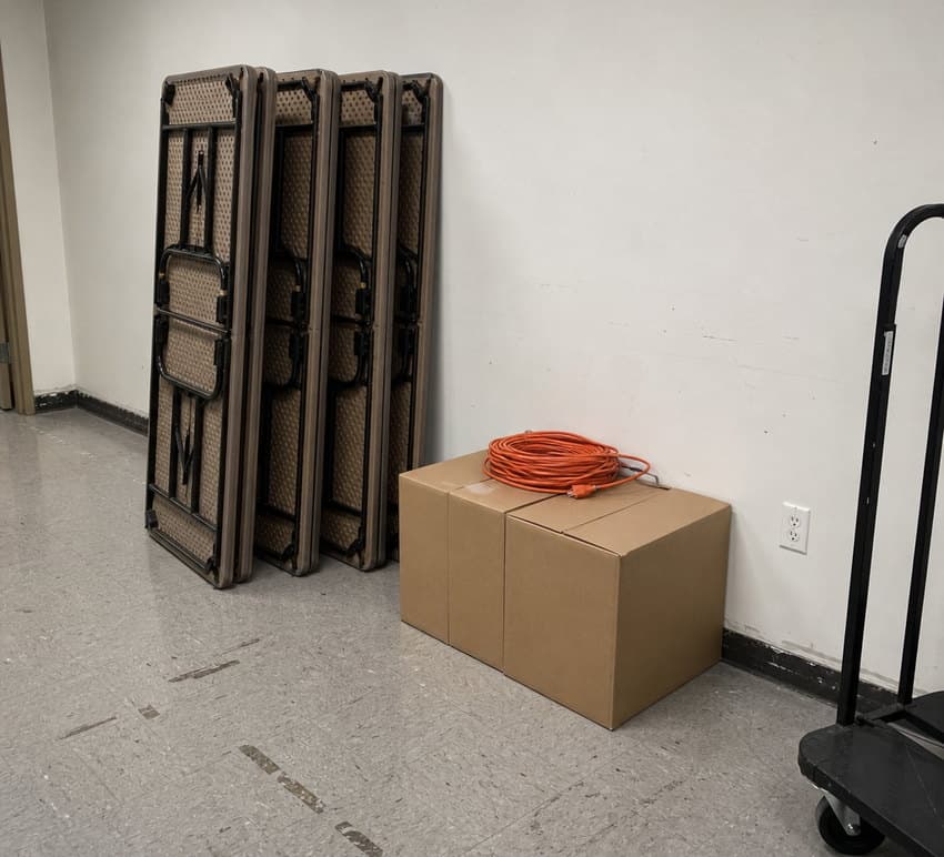 Folding tables stacked against a bare wall, plain cardboard boxes on the floor, a coil of orange extension cable on top.